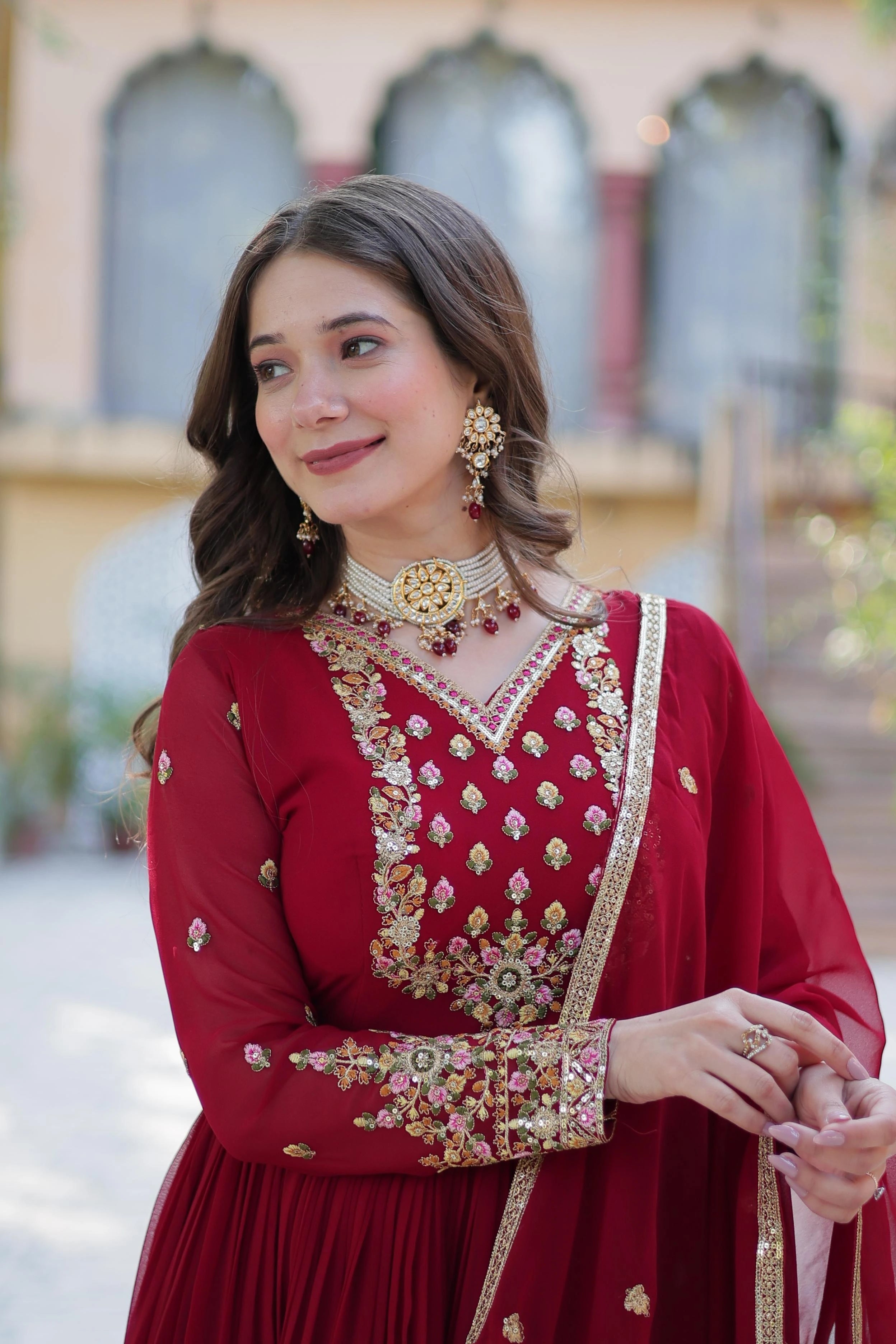Woman in traditional red outfit with gold embroidery and jewelry, standing outdoors.